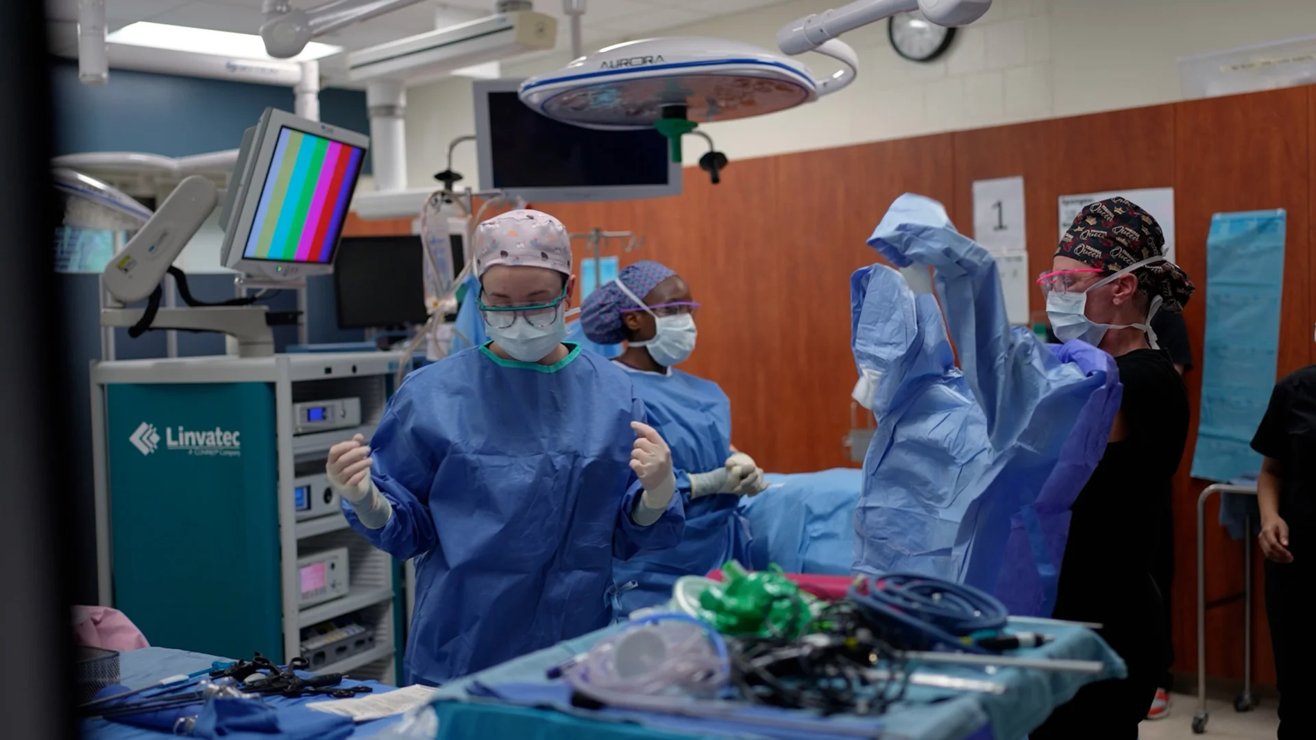 Students in a surgical technology training lab prepare for a procedure in a simulated operating room. Several individuals wear blue sterile gowns, masks, gloves, and protective eyewear while standing near surgical equipment, monitors, overhead operating lights, and a table of medical instruments.