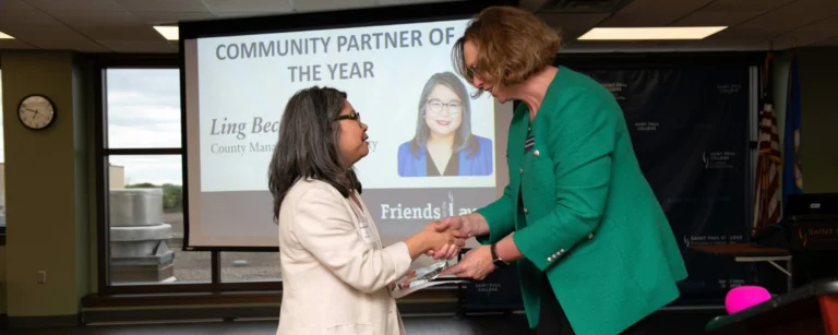 Ling Becker accepts a Community Partner of the Year award from President Peaslee during Friends on the Lawn. The two women shake hands in front of a projection screen displaying the award announcement.