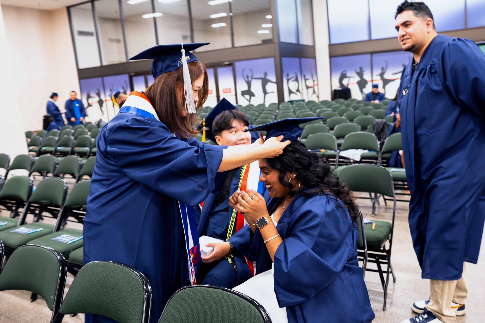 Students in blue graduation gowns celebrate together before a ceremony, one placing a cap on another's head.