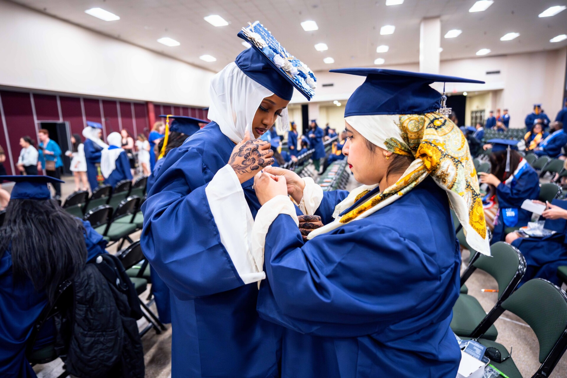 Two women in blue graduation gowns help each other with their outfits, in a crowded hall.
