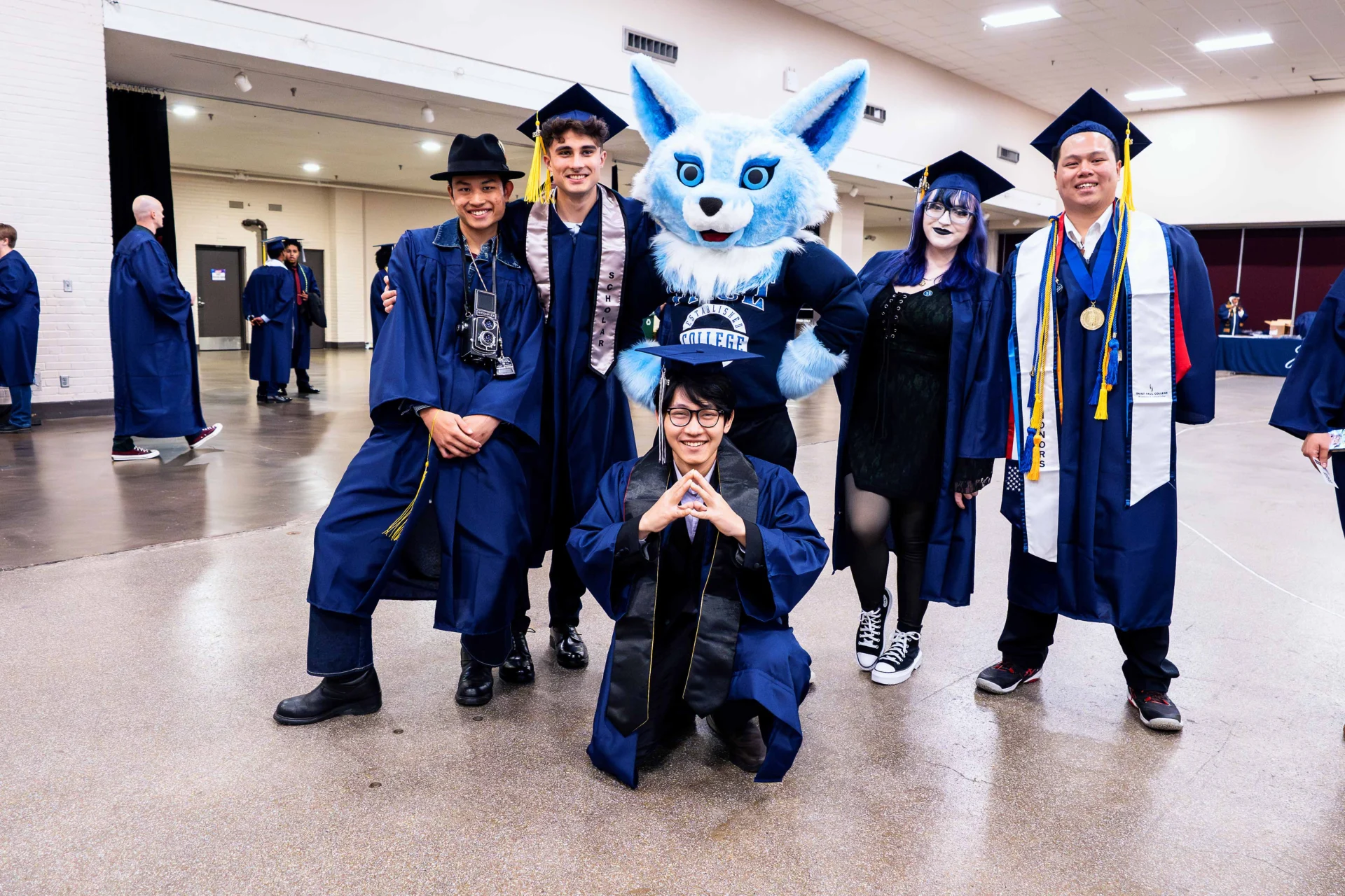 A group of graduates in blue caps and gowns posing together, including Sky the mascot and various accessories.