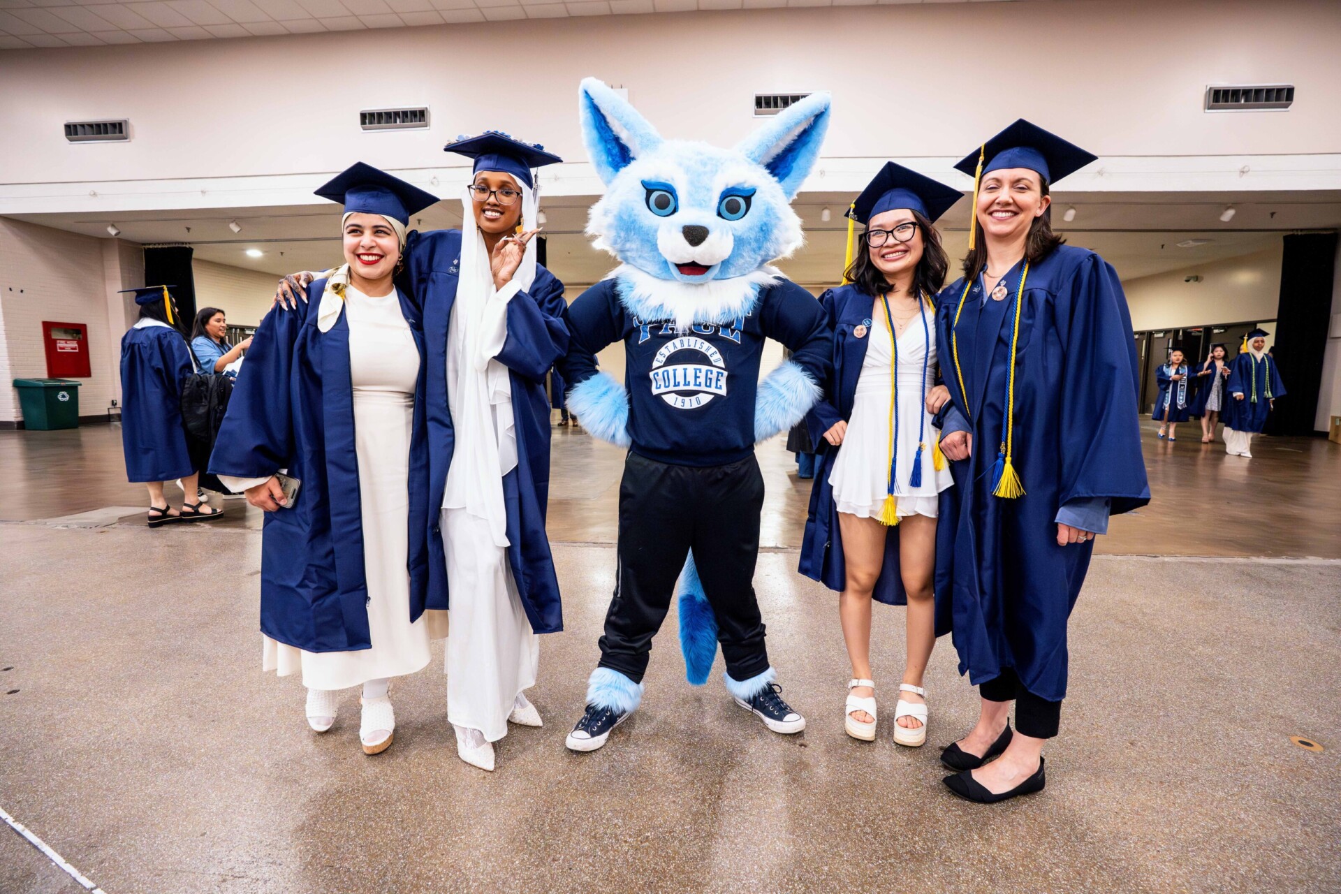 Four graduates in blue gowns pose with a blue fox mascot in a large indoor space.
