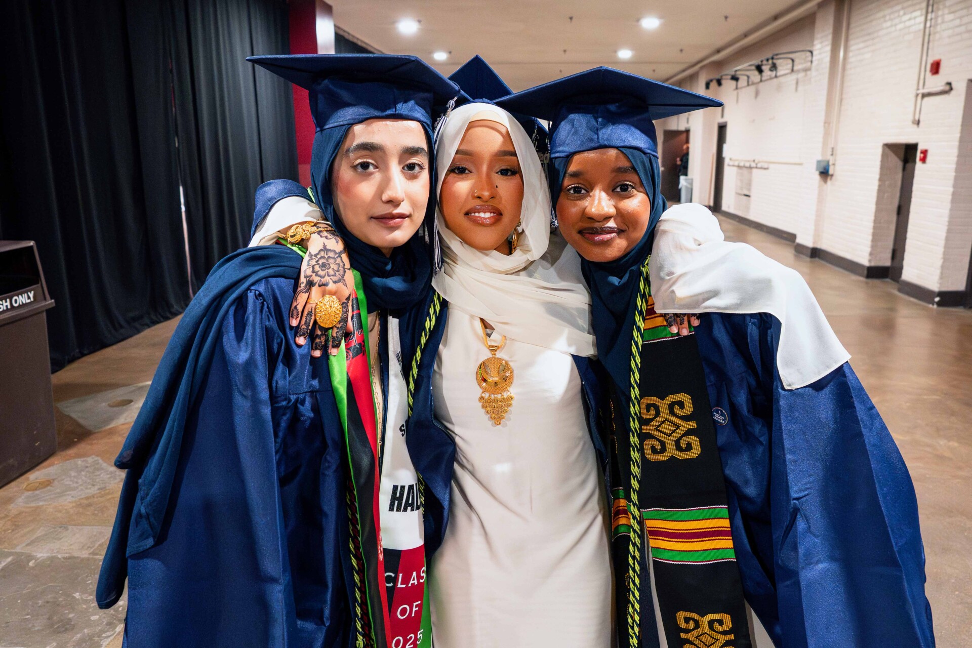 Three graduates in blue gowns and caps, hugging in celebration. One is in a white outfit with a gold necklace.