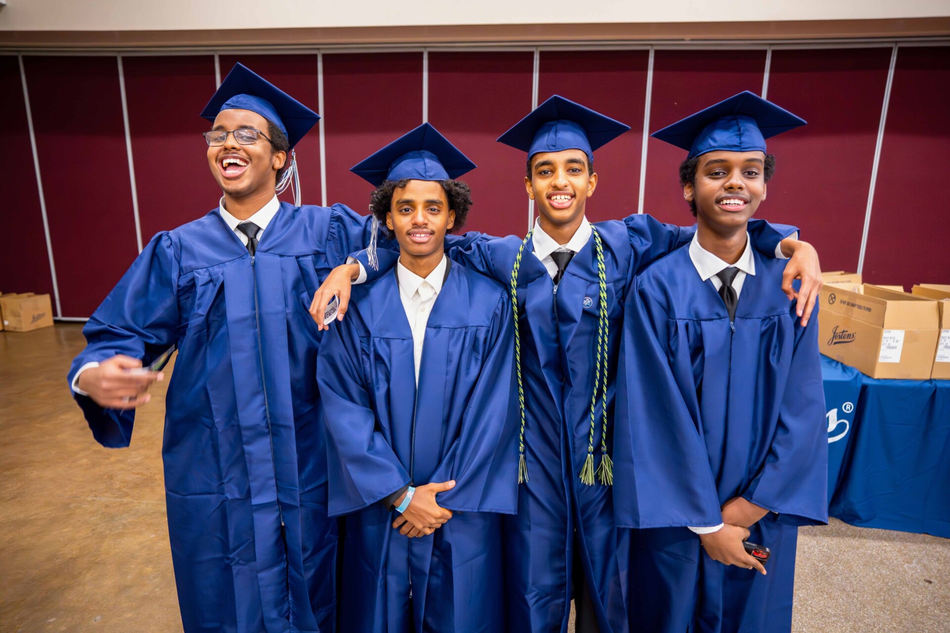 Four young men in blue graduation gowns and caps, smiling and posing together.