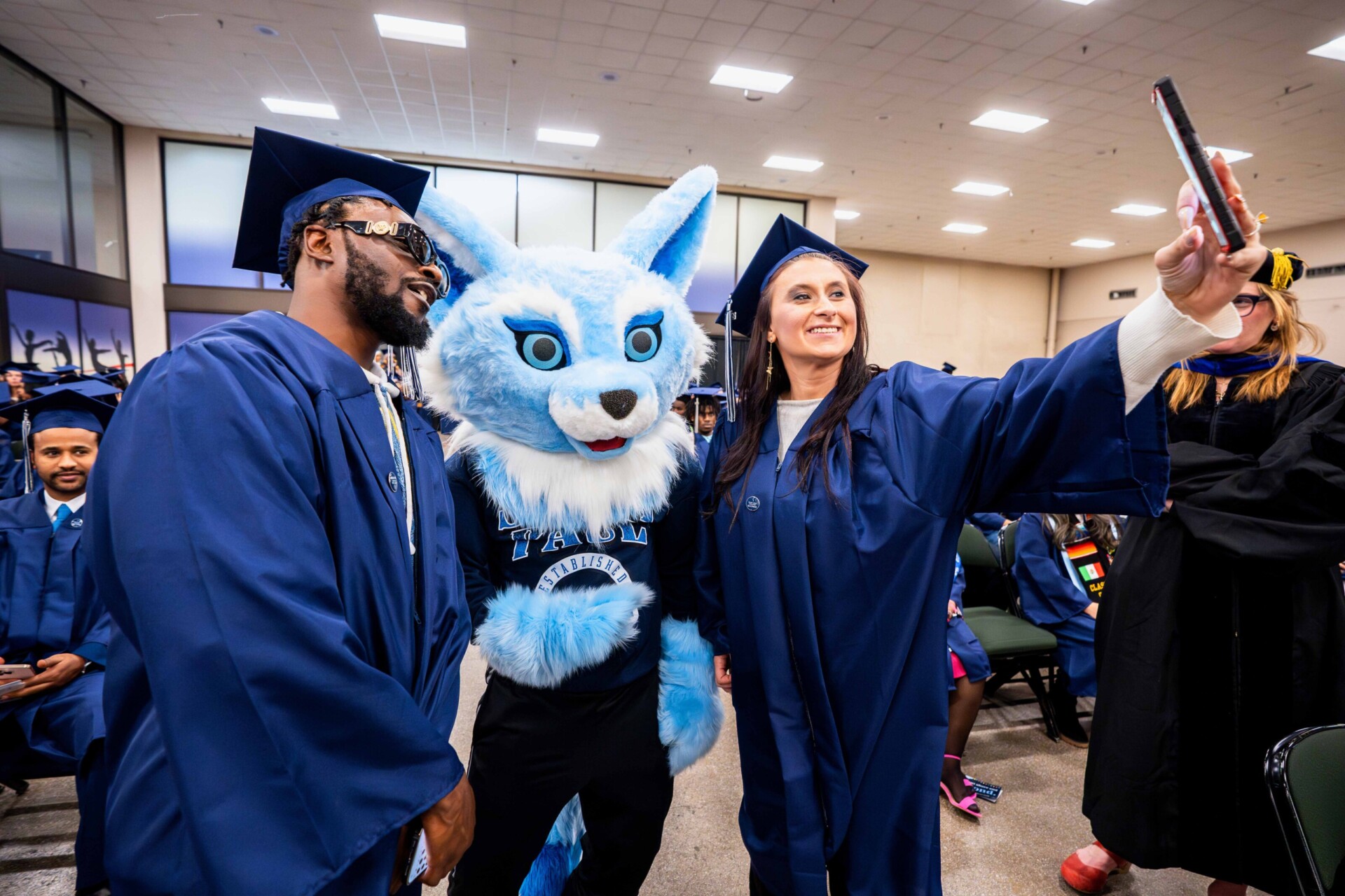 Two graduates in blue caps and gowns take a selfie with Sky the mascot at a graduation ceremony.