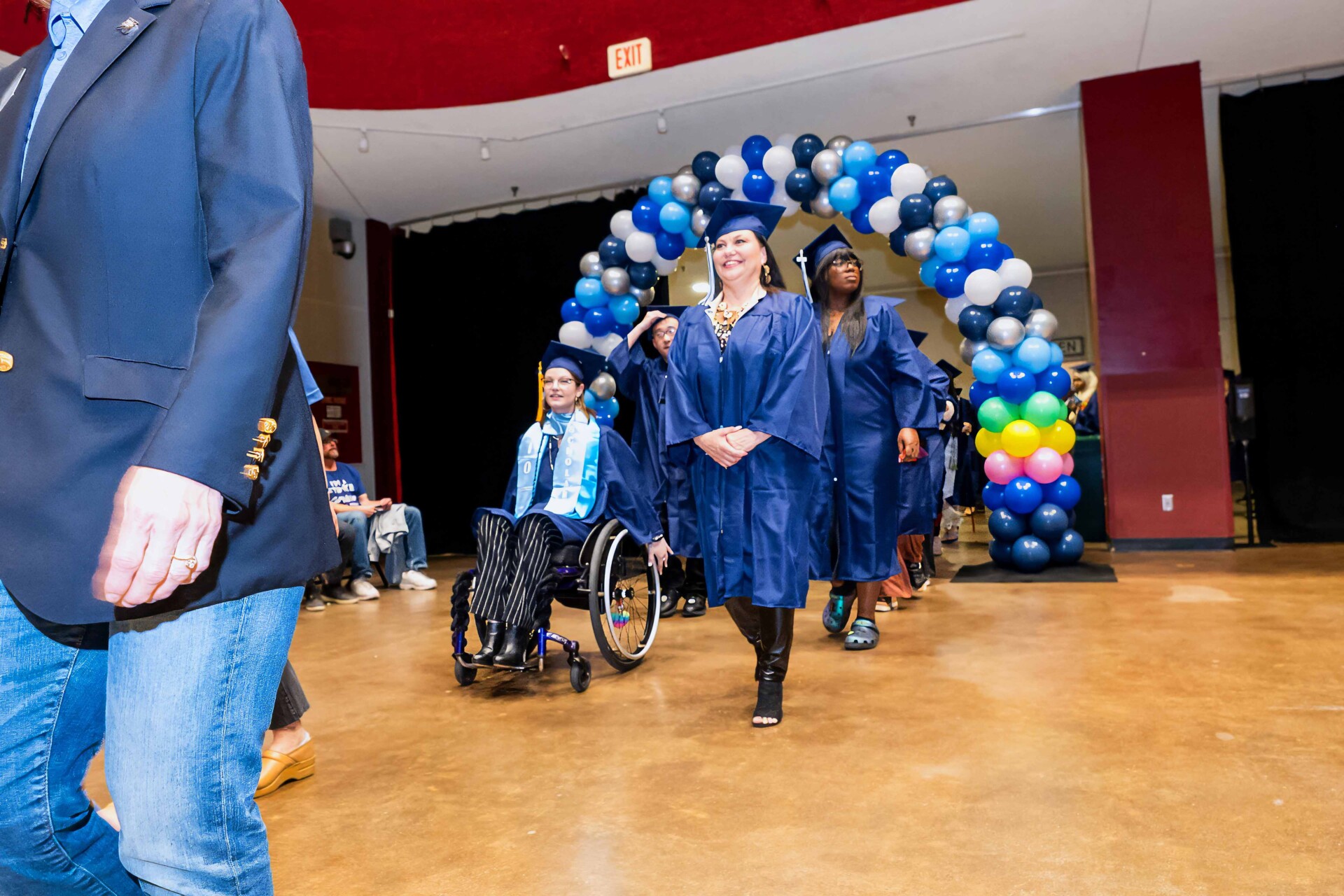 Graduates in blue caps and gowns walking through a balloon arch, with one in a wheelchair.