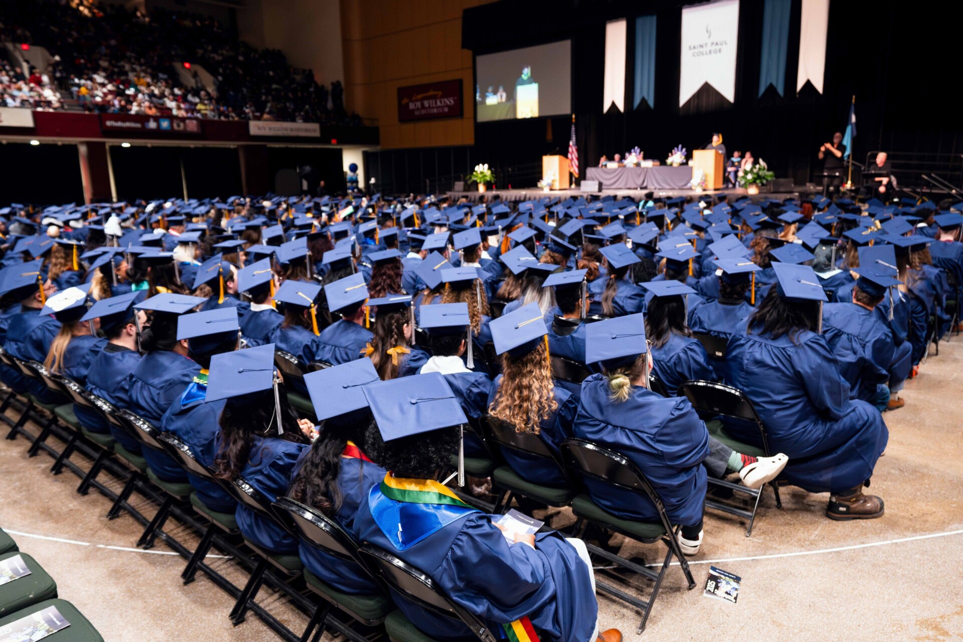 A packed graduation ceremony with many graduates in blue gowns and caps. The stage has speakers. Chairs are arranged.