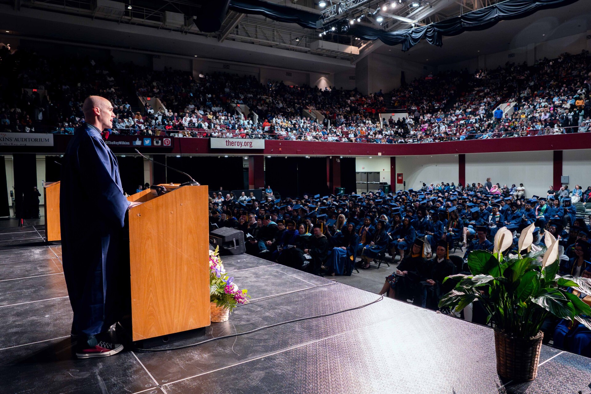 Michael Blackwood in a graduation gown stands at a podium, addressing a large audience of graduates, all in caps and gowns.