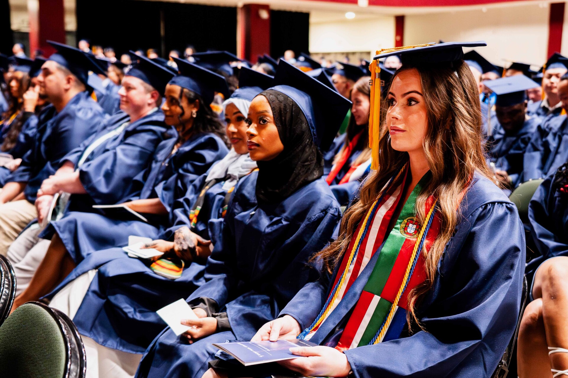 Graduates in caps and gowns sit together during a graduation ceremony, focused and attentive.