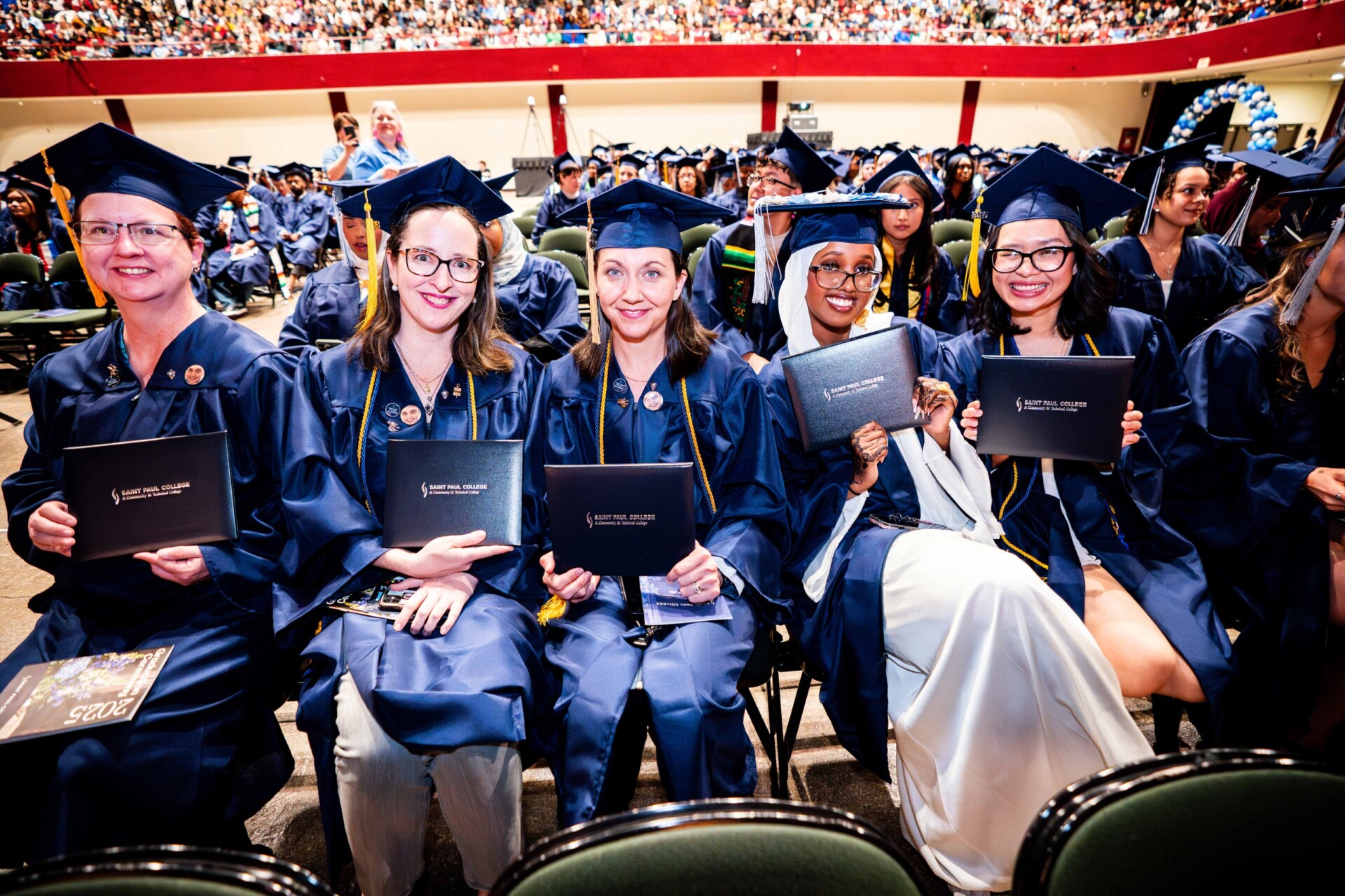 Five graduates in blue gowns and caps holding diplomas, smiling at the camera.