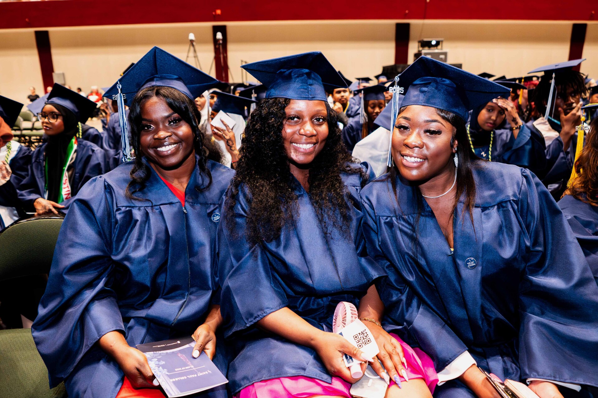 Three smiling graduates in blue gowns and caps, sitting together at a ceremony.