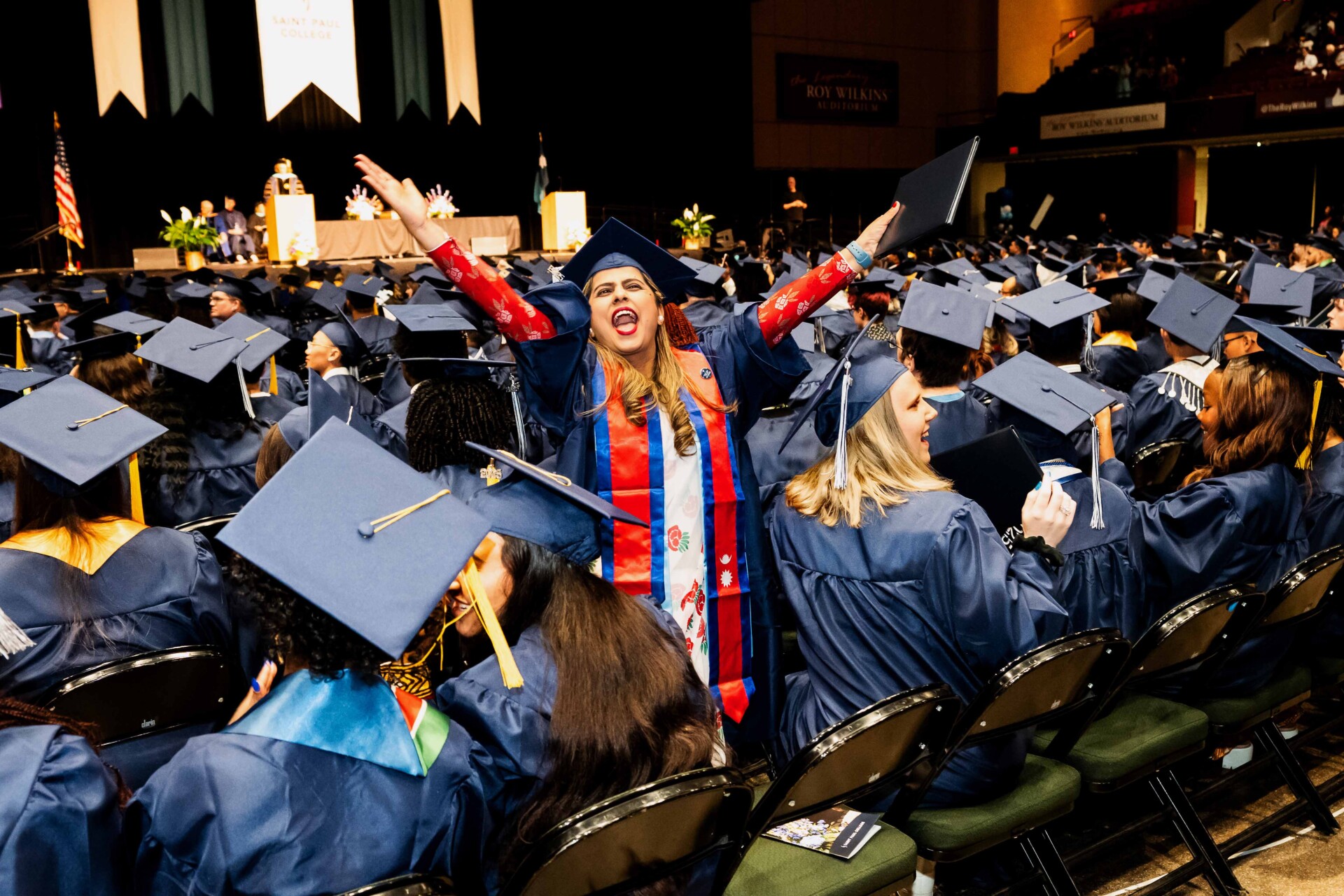 A joyous graduate in a blue cap and gown celebrates with arms raised at a graduation ceremony.