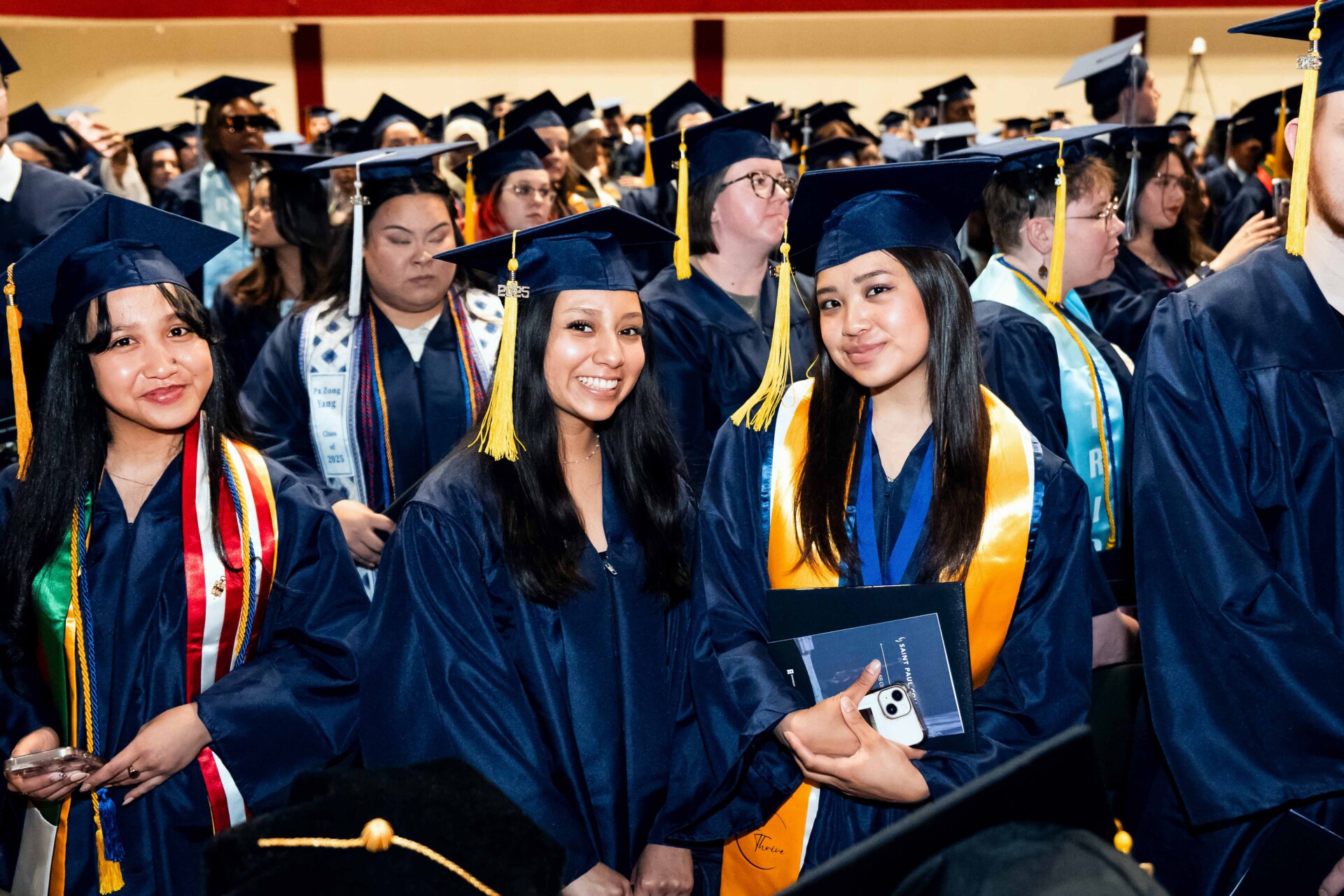 Three graduates wearing blue gowns, caps, and sashes, smiling at a graduation ceremony.