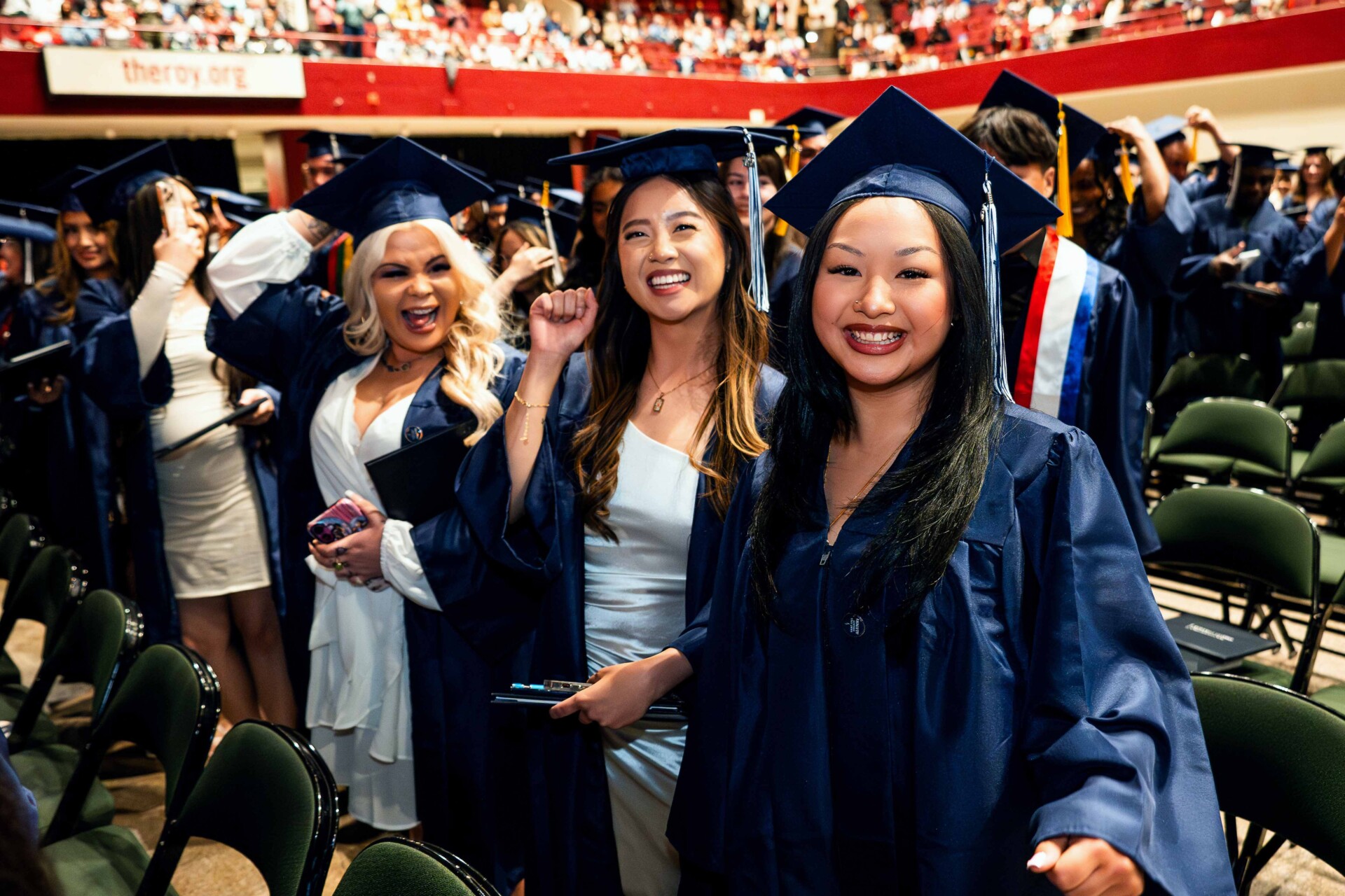 Three smiling graduates in blue caps and gowns celebrate at a graduation ceremony.