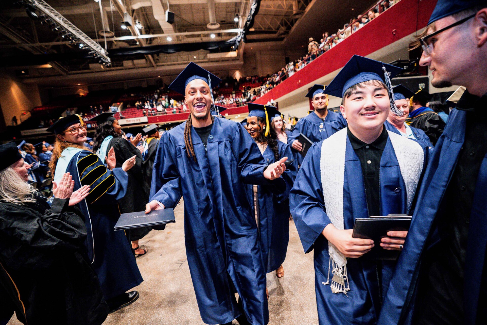 Graduates in blue caps and gowns celebrate on stage, holding diplomas.
