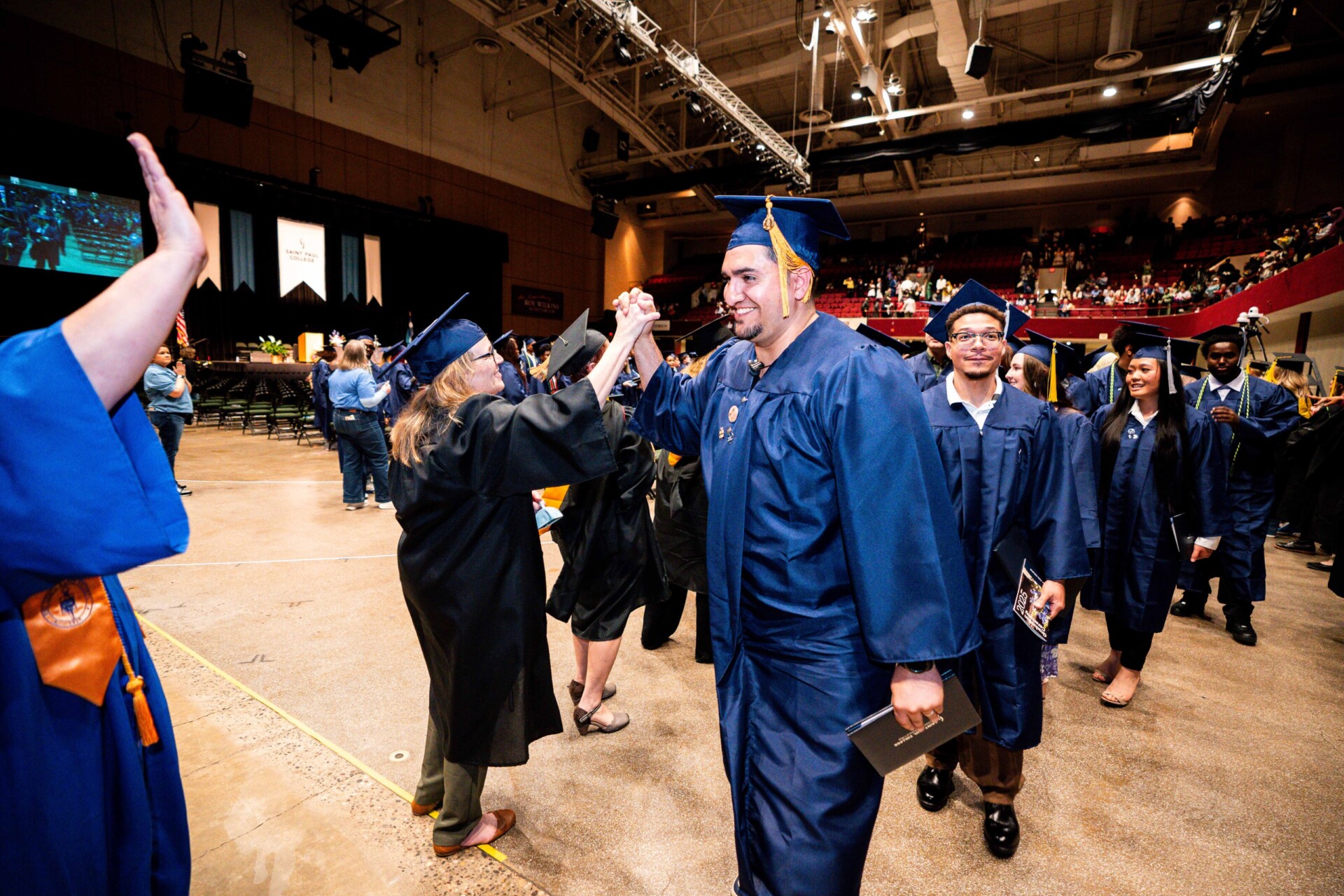 Graduates in blue gowns celebrating and high-fiving during a graduation ceremony.