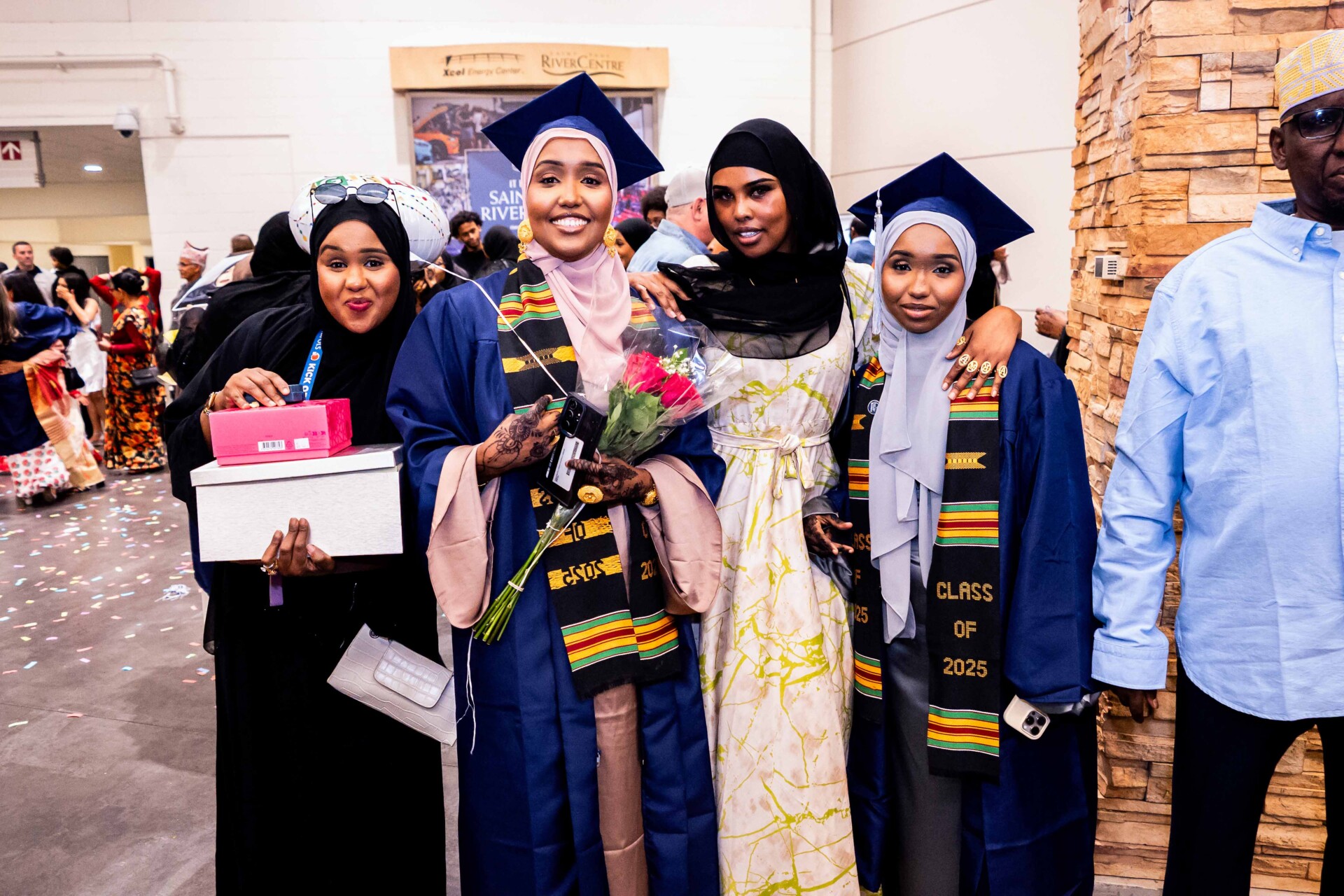 Four young women celebrate graduation, wearing caps and gowns. One holds flowers and gifts.