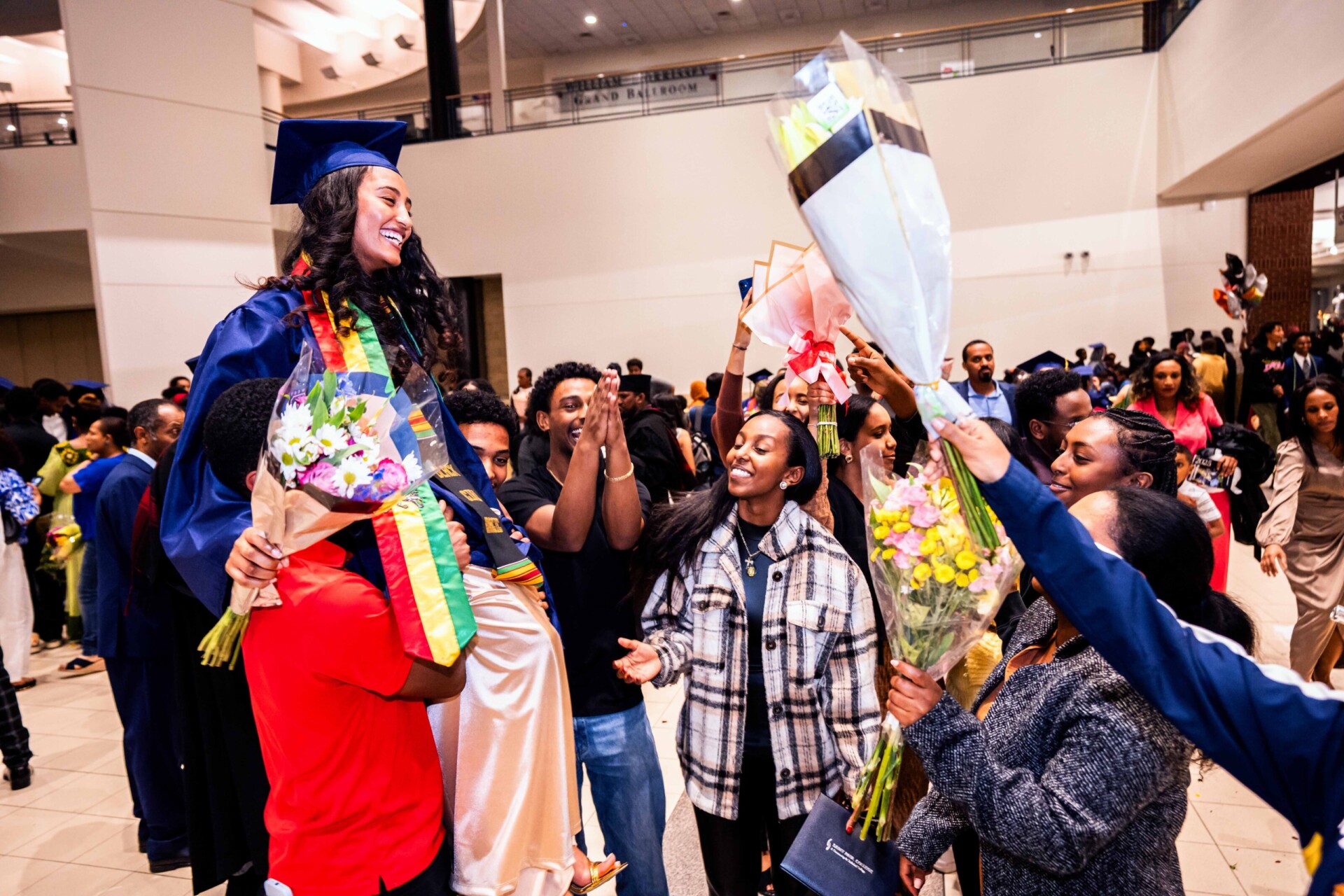 A graduate in a blue cap and gown smiles while being lifted by friends, surrounded by flowers and celebration.