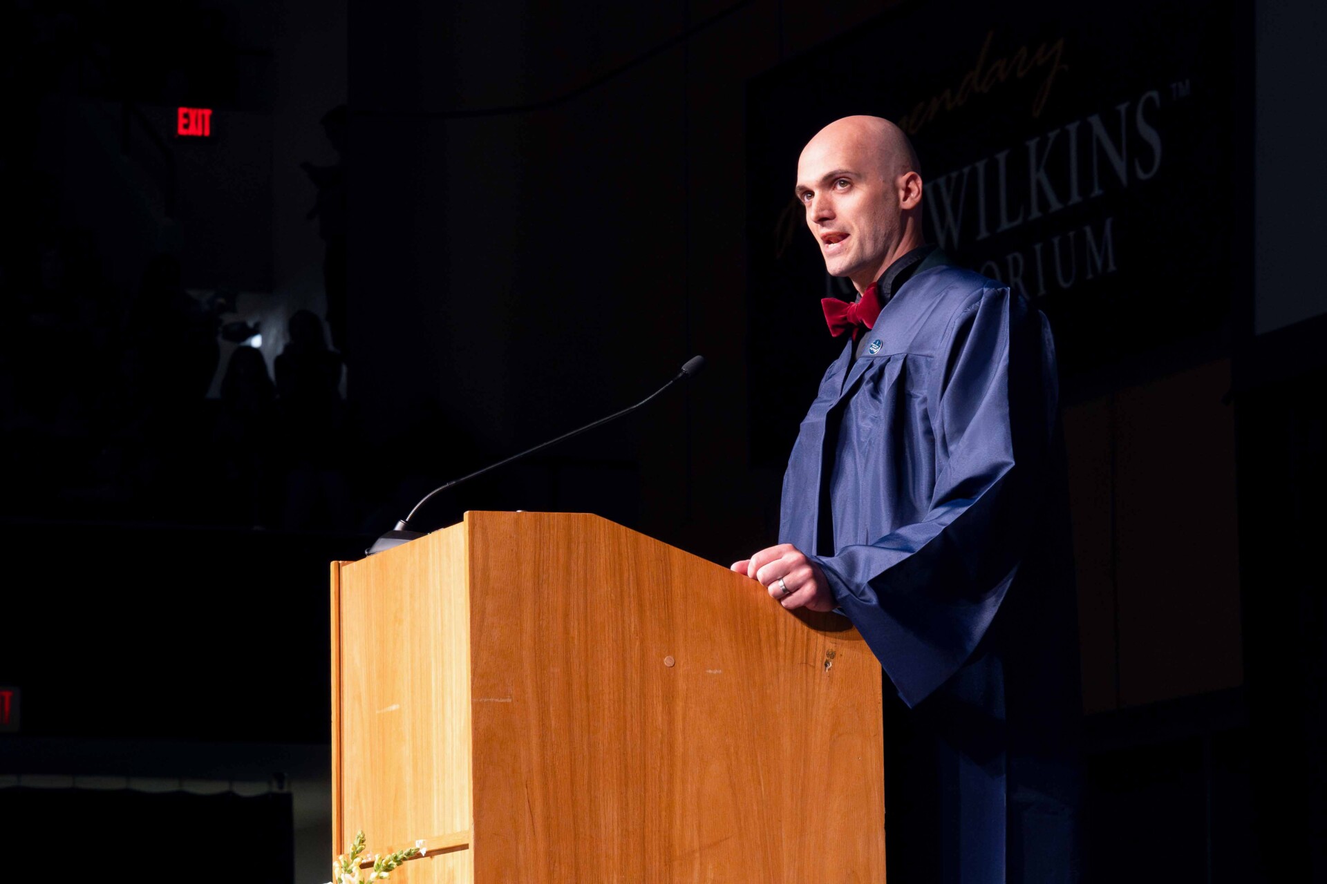 Michael Blackwood in a graduation gown and bow tie stands at a wooden podium.