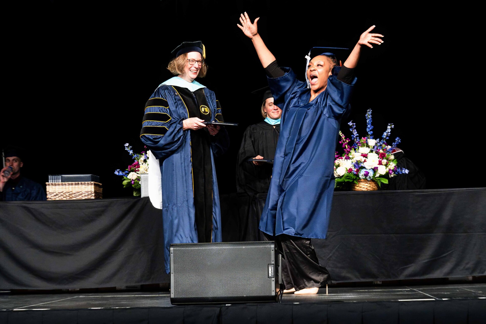 A graduate in a blue gown celebrates joyfully on stage, receiving a diploma from President Peaslee