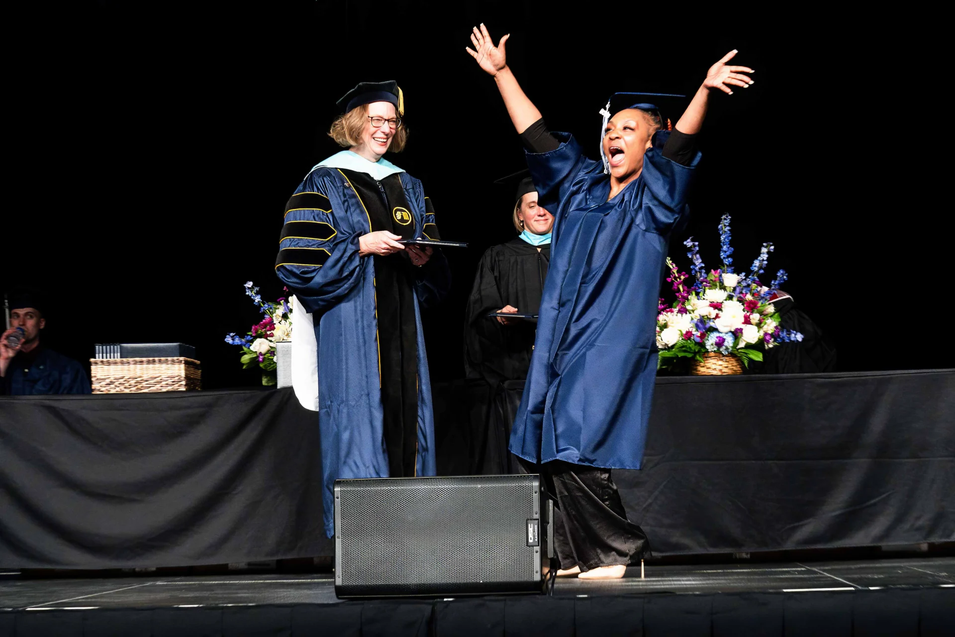 A graduate in a blue gown celebrates joyfully on stage, receiving a diploma from President Peaslee