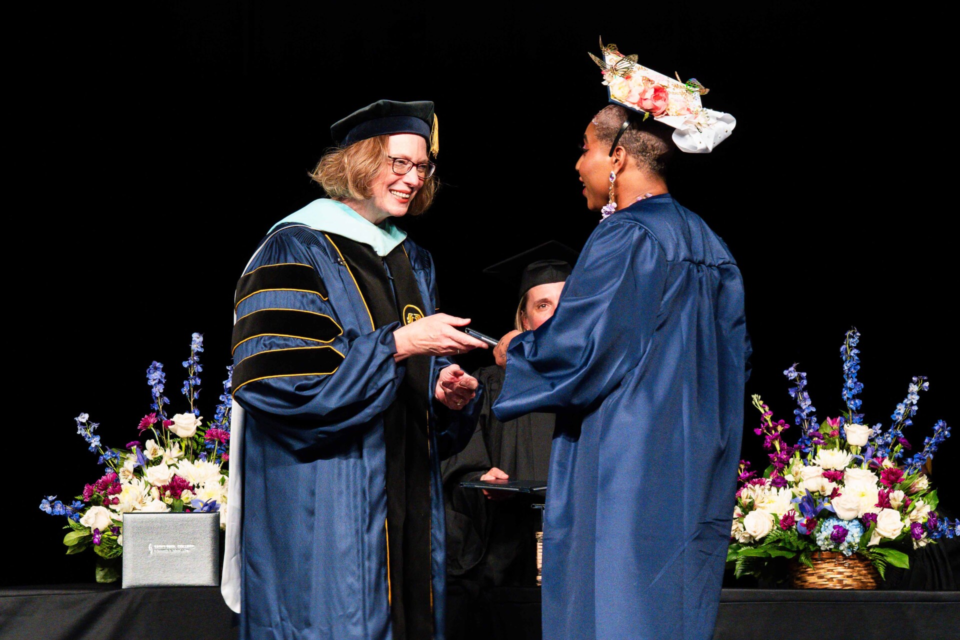 A graduate in a blue gown is receiving a diploma from President Peaslee in academic regalia. Flowers decorate the stage.