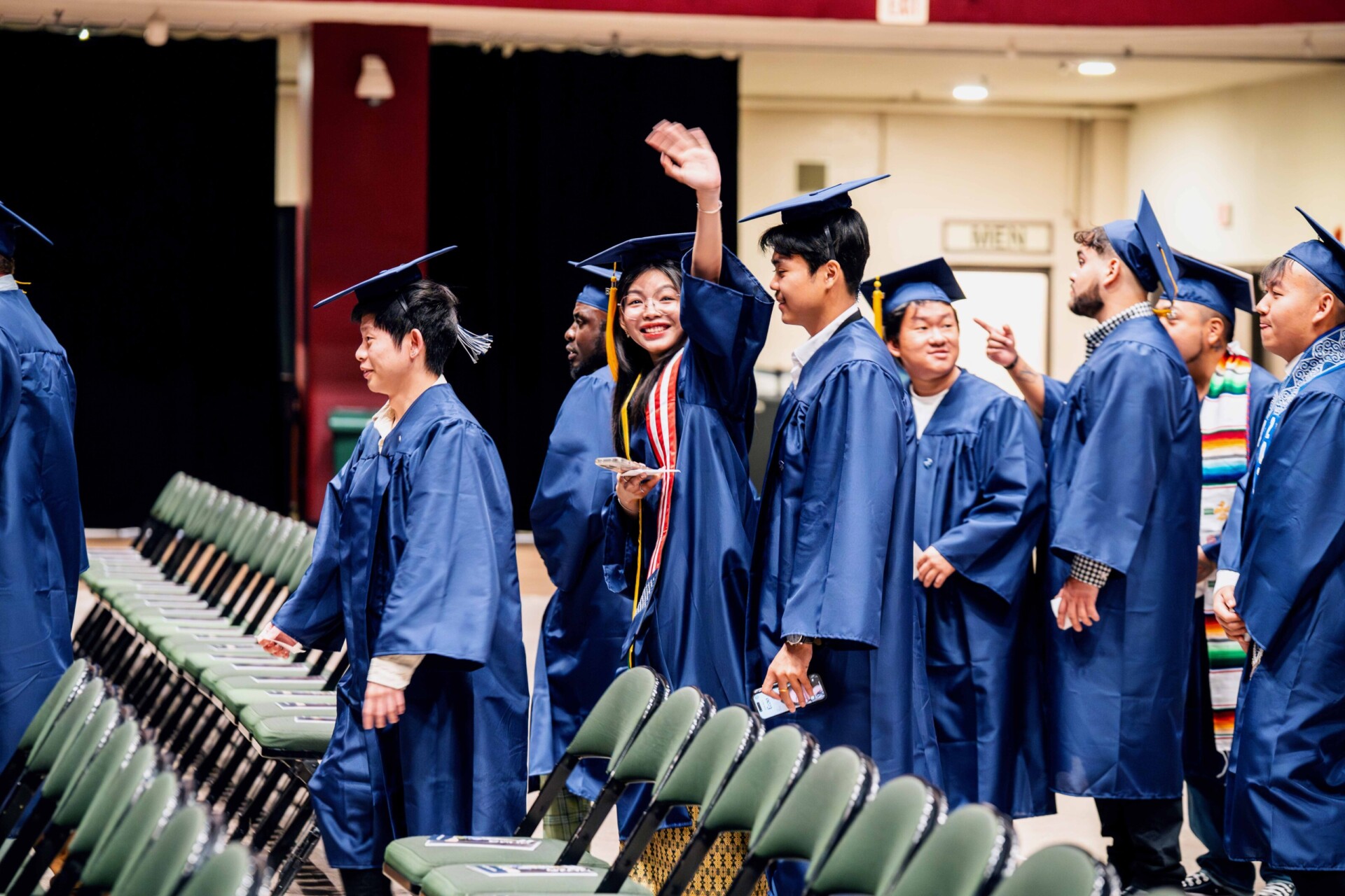 Graduates in blue caps and gowns walking and waving, smiling in a ceremony setting with green chairs.