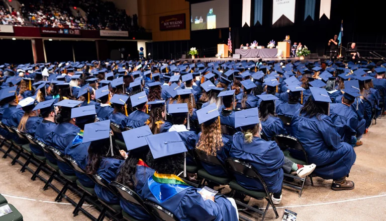 A packed graduation ceremony with many graduates in blue gowns and caps. The stage has speakers. Chairs are arranged.