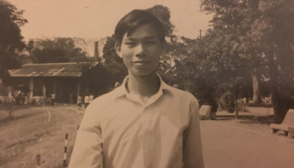 Francois Nguyen as a young man in a white shirt smiles, standing outdoors with trees and a building in the background. Photo is sepia.