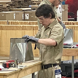 Dylan Olson in a beige shirt and gloves assembling metal parts at a workshop table. 