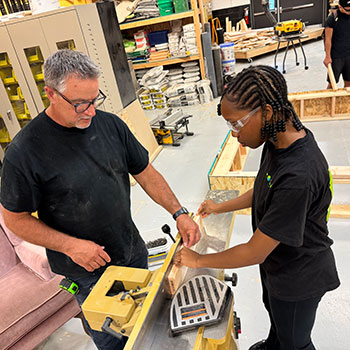 Tom Leiskau and a student in black clothes work together on a woodworking project, using a table saw.