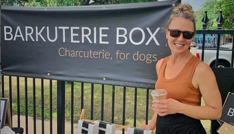 Rebecca Jackson holding a drink stands by a booth with a sign for Barkuterie Box, offering dog charcuterie.