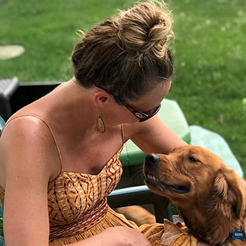 Rebecca Jackson  in a patterned dress, smiling and petting a golden retriever in a green outdoor setting. 