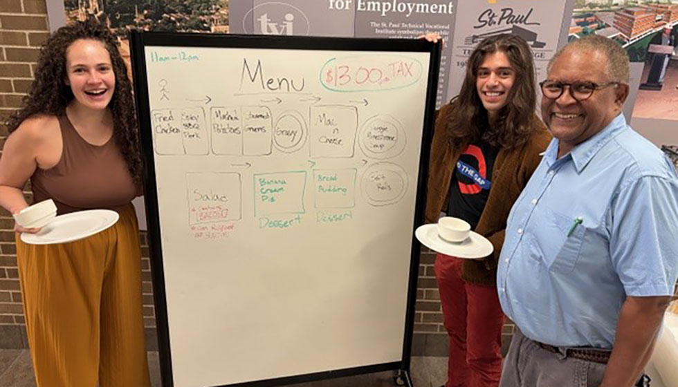 Three people standing next to a menu board, smiling. One holds a plate, another holds a cup.