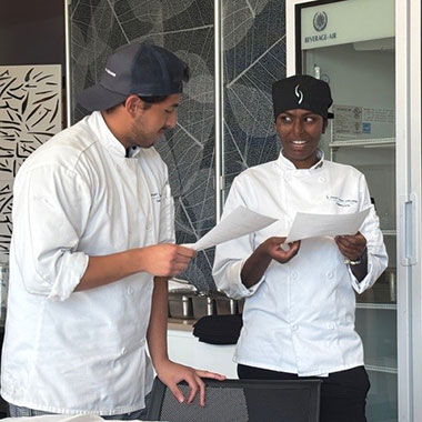 Two chefs talking while holding paper notes in a modern kitchen.