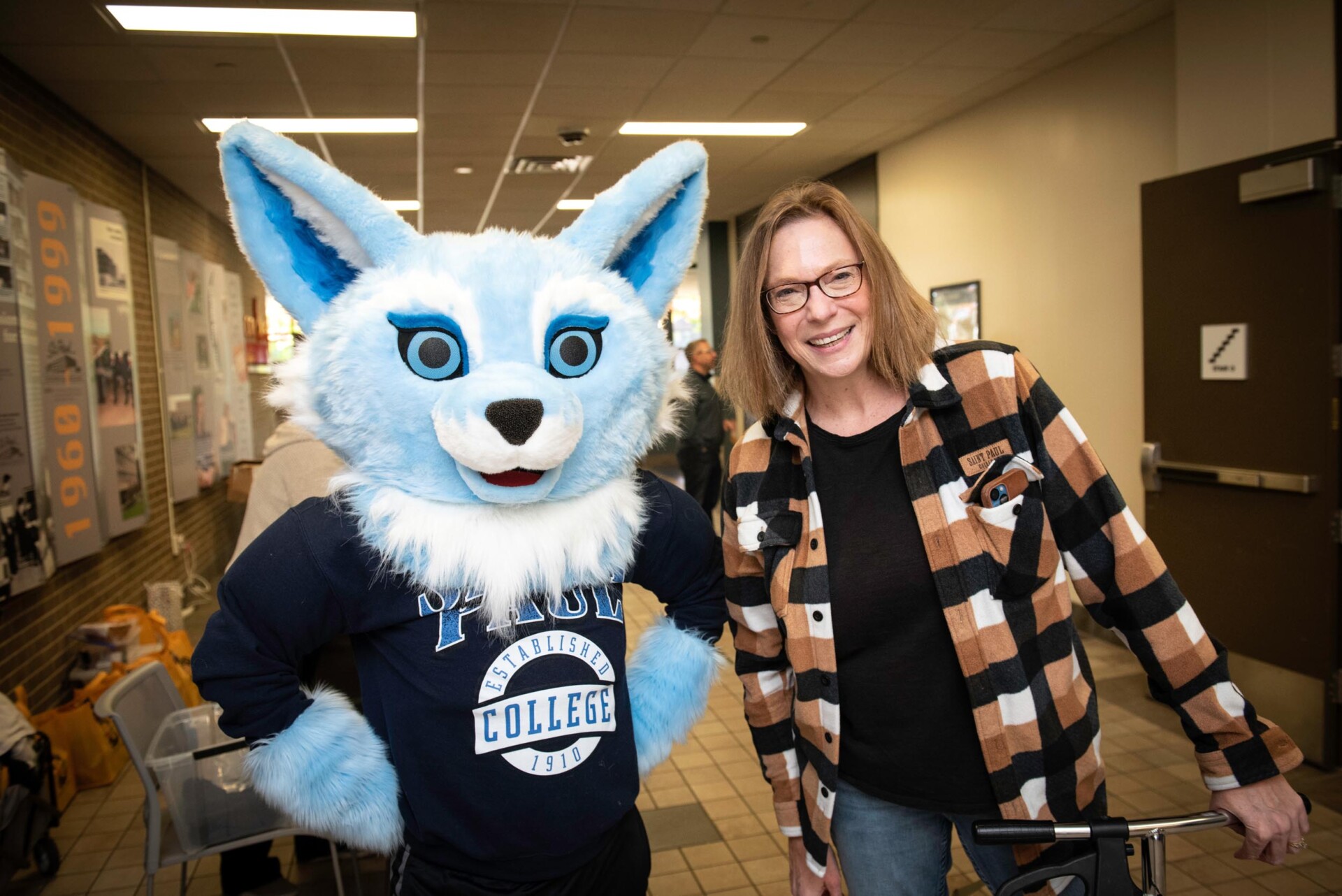 Dee Dee Peaslee stand next to Sky the fox mascot wearing a Saint Paul College shirt. They are in a hallway with informational posters on the wall and a doorway in the background.