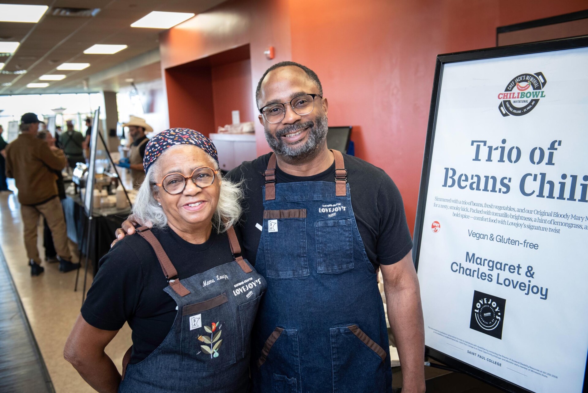 Two people wearing matching dark aprons with “Lovejoy’s” branding stand next to a large sign that reads “Trio of Beans Chili” with details about the dish being vegan and gluten-free, credited to Margaret & Charles Lovejoy. The background shows a room with tables and people.