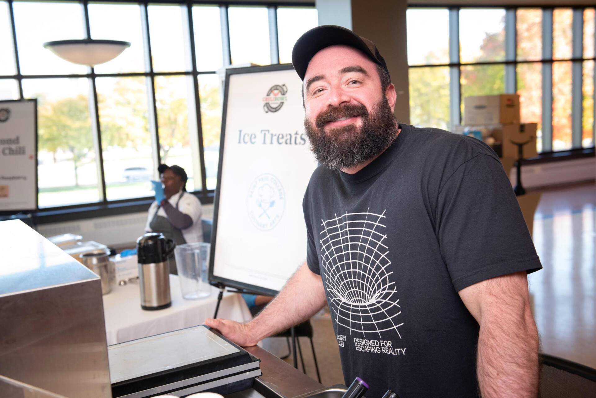 A person wearing a black T-shirt with a geometric design stands near a counter with equipment. Behind them is a sign for “Ice Treats” and another person in culinary attire at a table with a blender and coffee dispenser.