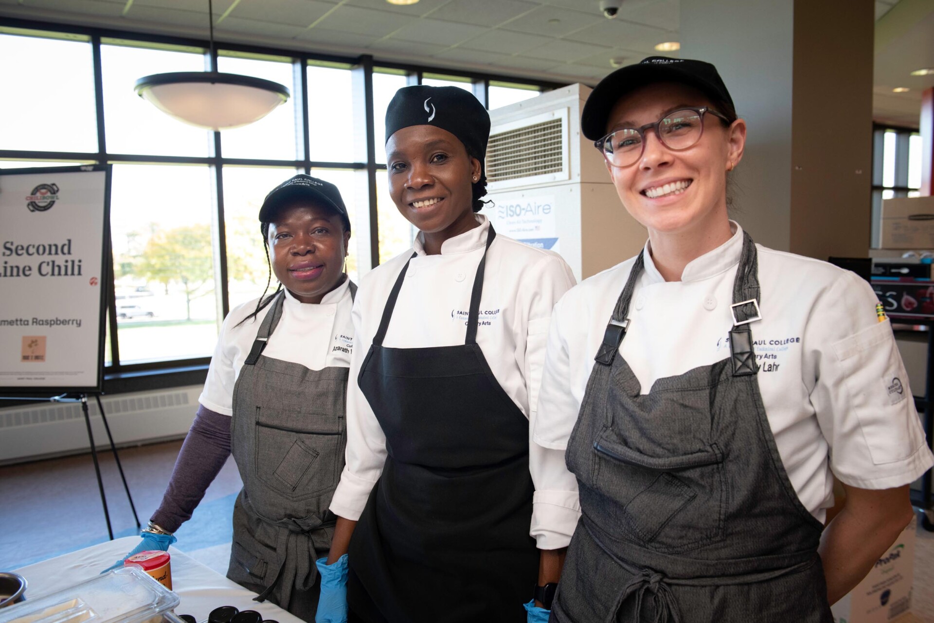 Three students in white chef coats and gray aprons stand behind a table with culinary items. A sign in the background reads “Second Line Chili” with the name Demetta Rosenberg. Large windows provide natural light.