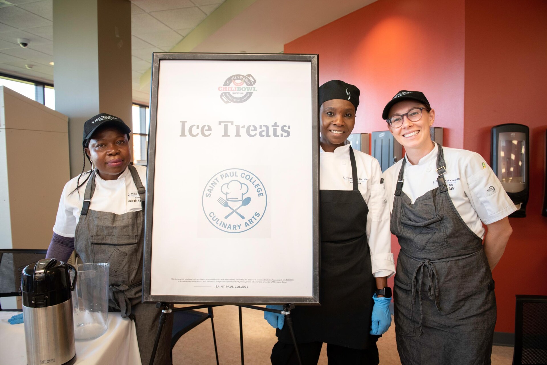 Three students dressed in white chef coats and gray aprons stand beside a sign that reads “Ice Treats” with the Saint Paul College Culinary Arts logo. A blender and coffee dispenser are visible on the table in front of them.