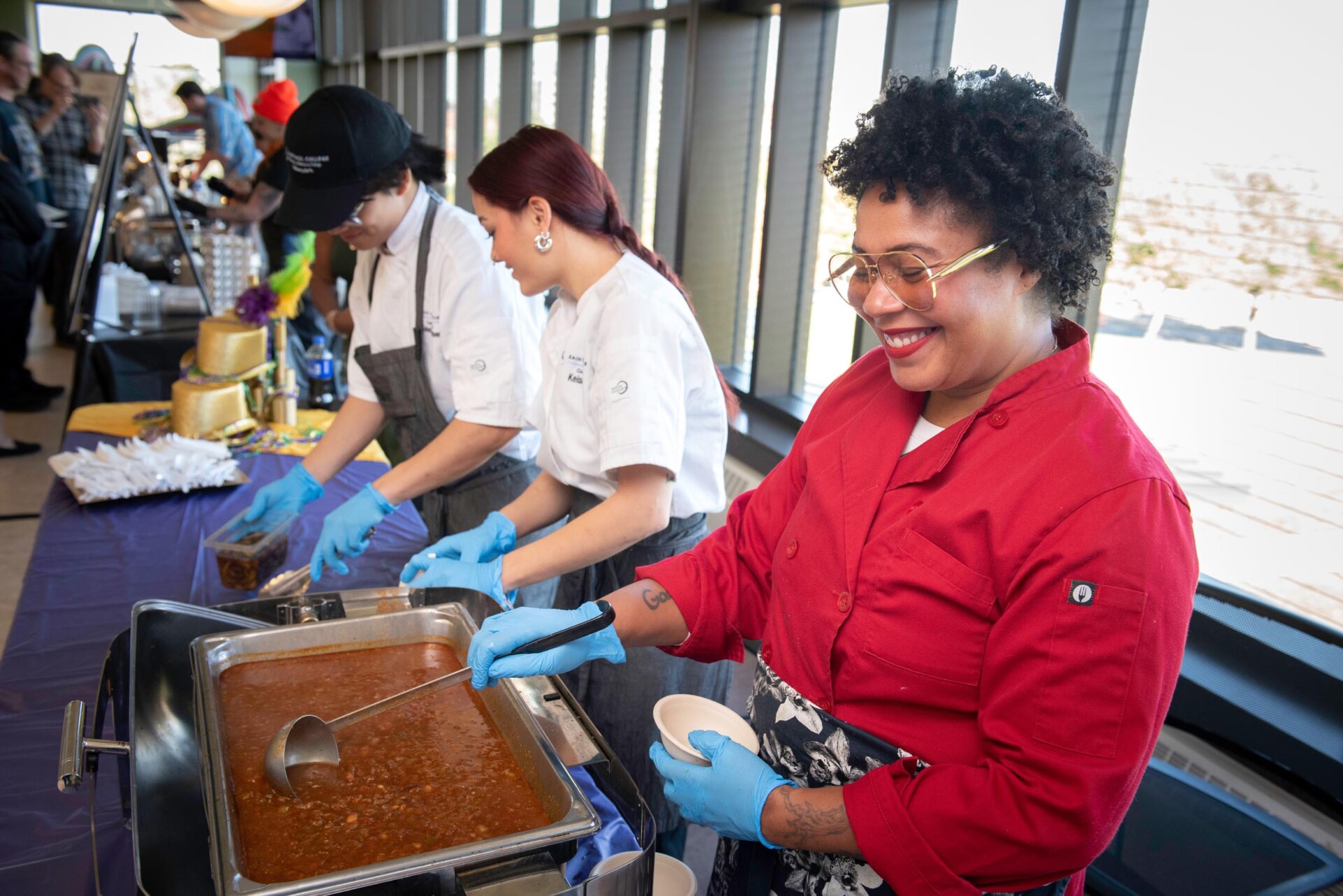 A person in a red chef coat serves chili from a large stainless-steel pan into a small white bowl, while two individuals in white chef coats and gray aprons prepare food beside them. The setup is on a long table covered with a dark cloth near large windows.