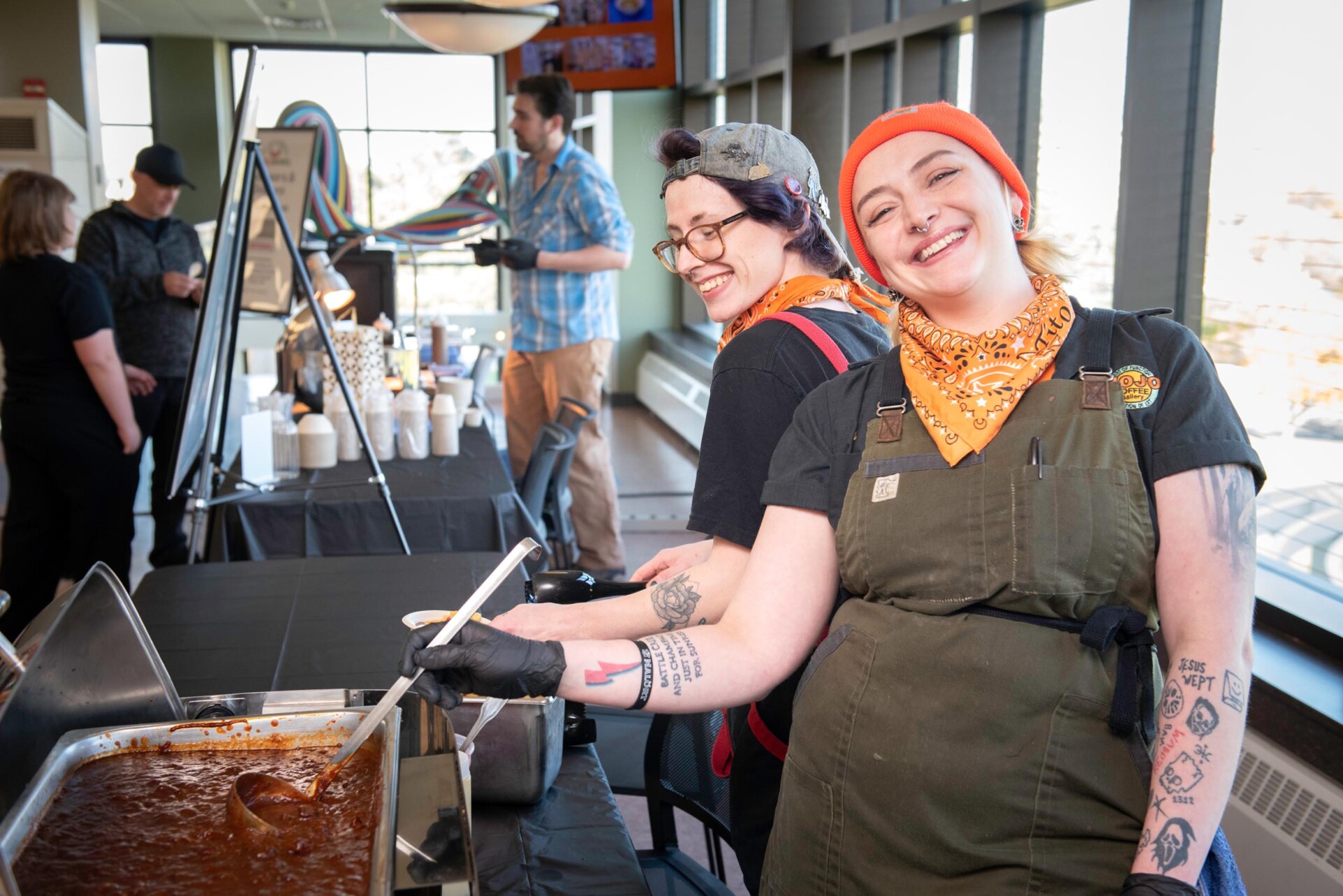Two individuals wearing black shirts, orange bandanas, and dark aprons stand behind a serving station with a large metal pan of chili. Condiment bottles are placed on the counter in front of them, and colorful autumn trees are visible through the windows.