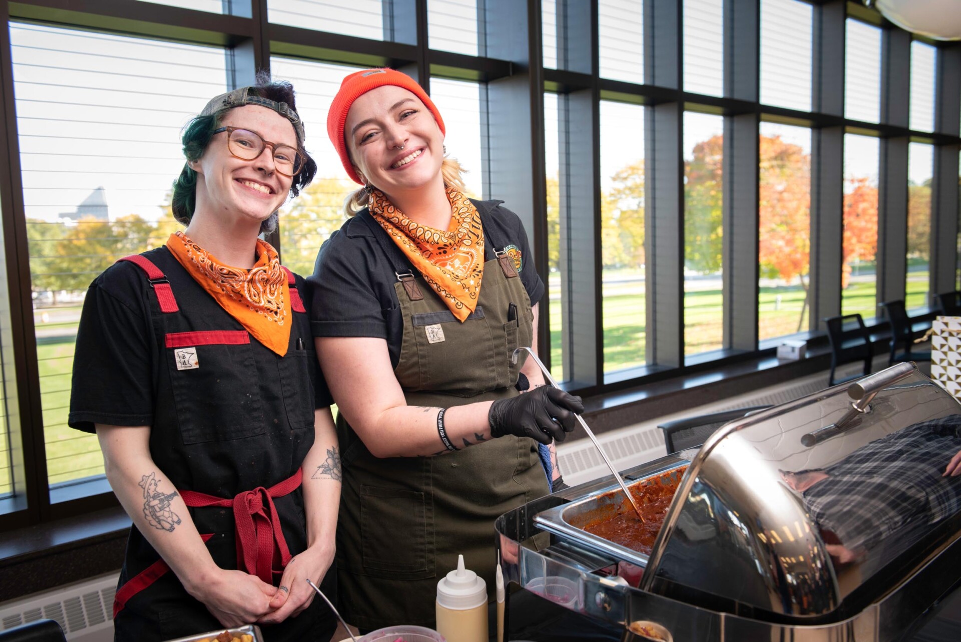 A person wearing a black shirt and orange bandana stands near a large pan of chili with a ladle inside, while another individual in similar attire is positioned slightly behind. The background shows a long table with various food items and people interacting.