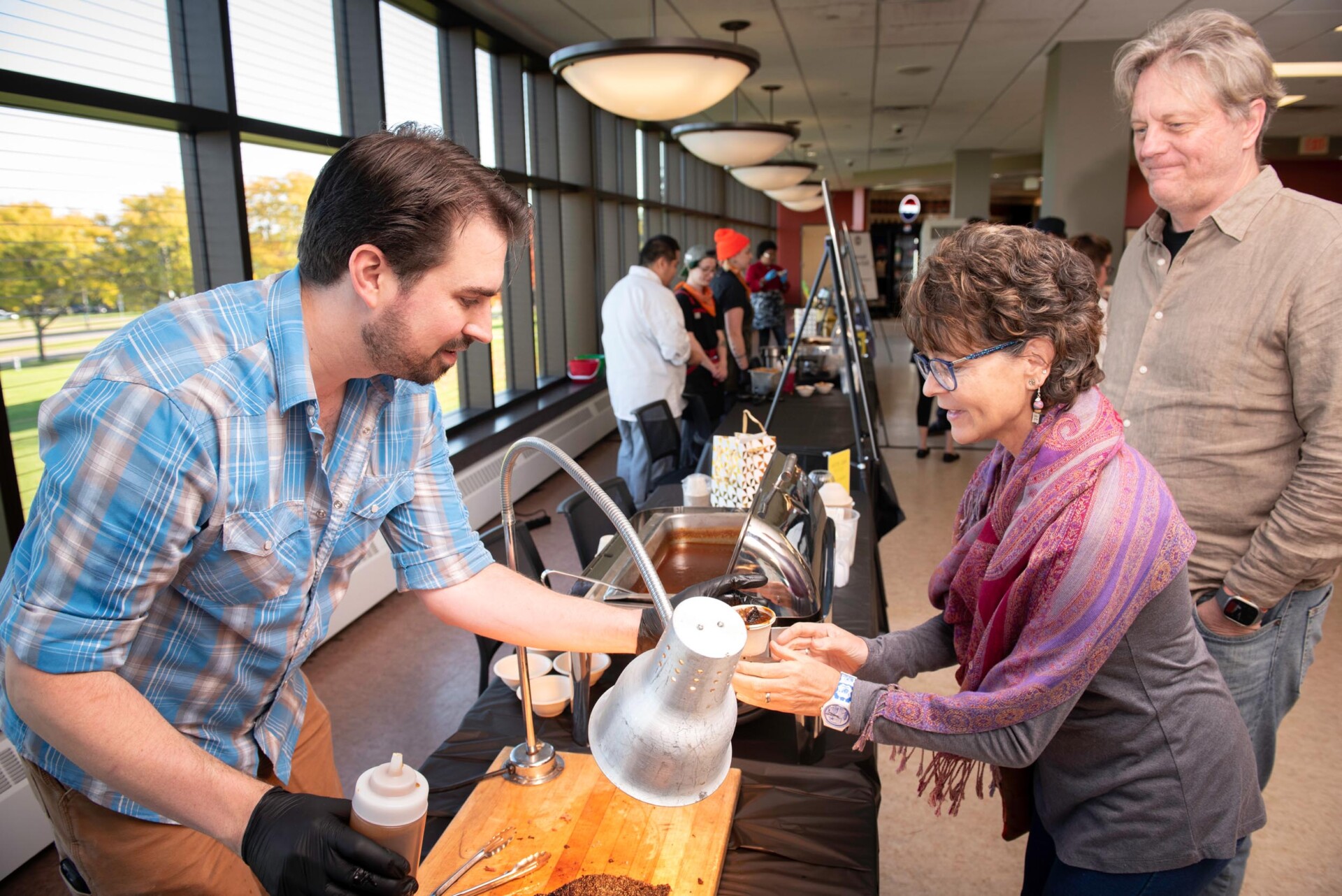 A person in a plaid shirt pours liquid from a kettle into a cup held by another individual wearing a purple scarf. A wooden board with a lamp and condiment bottle is on the table, and other food stations and people are visible in the background.