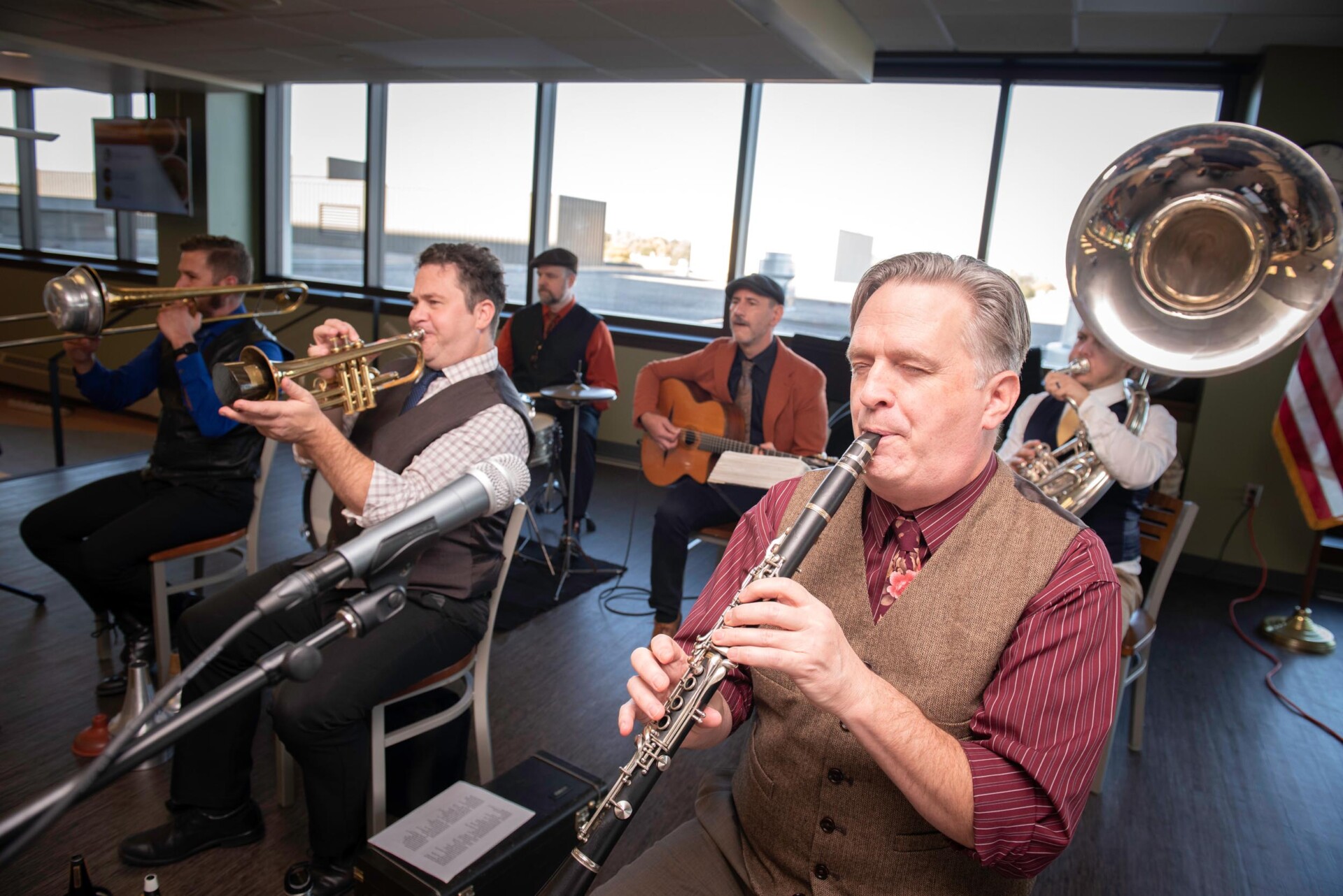 A band performs indoors near large windows. Visible instruments include a clarinet, trumpet, guitar, and sousaphone. Microphones and sheet music are set up in front of the musicians