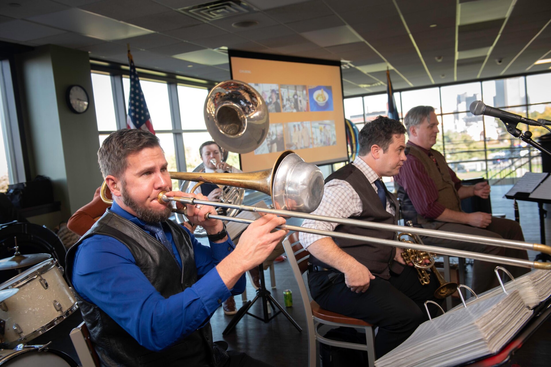 A musician in a blue shirt plays a trombone while seated among other band members holding brass instruments. A drum set and microphones are visible in the foreground, and a projection screen with images is displayed in the background near large windows
