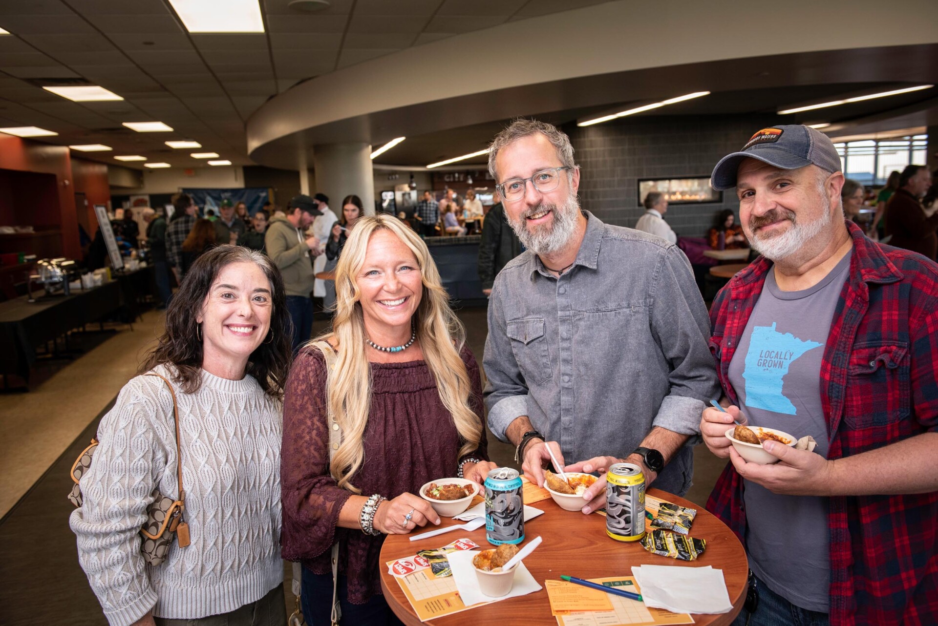 Four people stand around a tall round table holding bowls of chili and canned beverages. Event flyers and napkins are spread on the table, and the background shows a crowded indoor space with food stations.