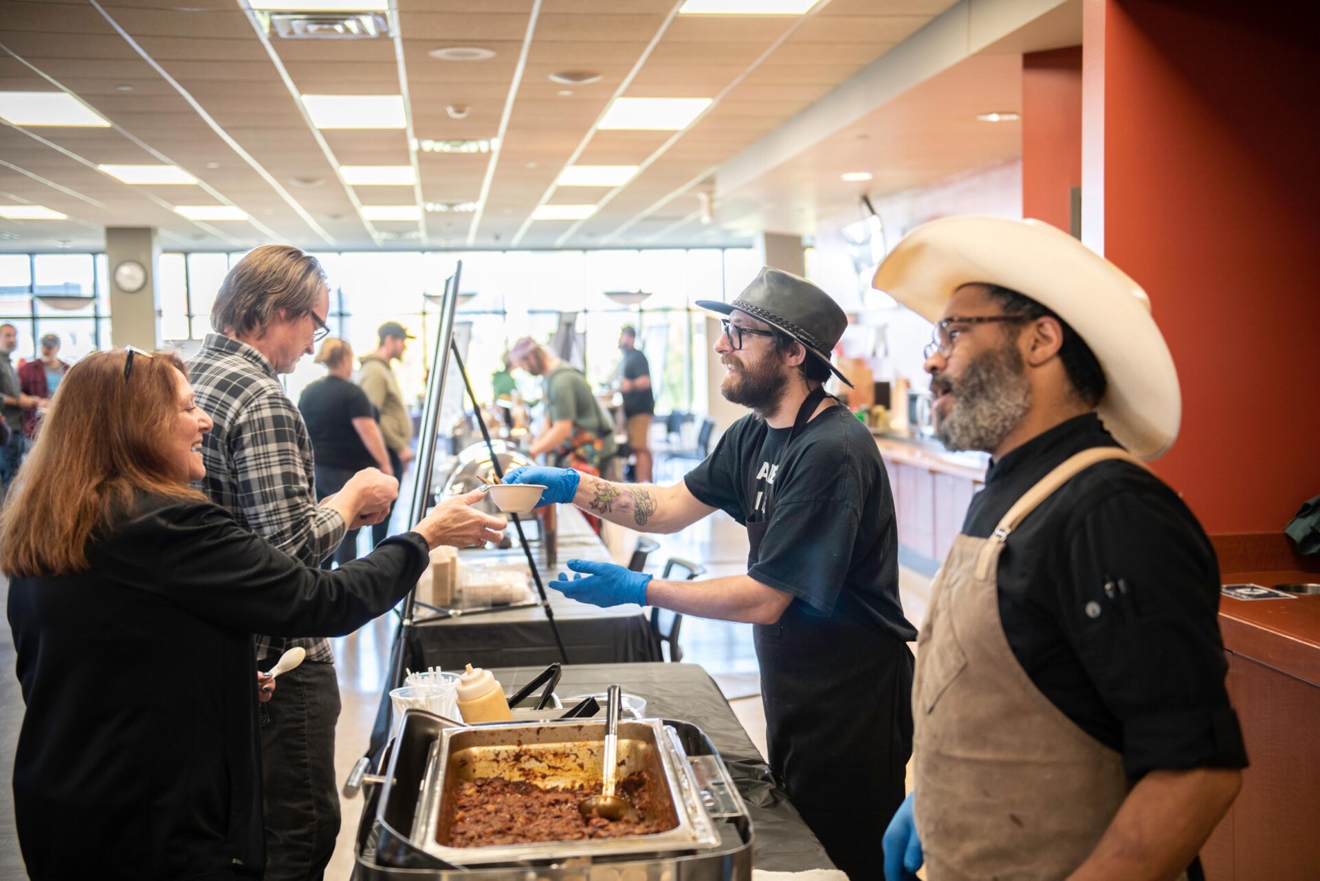 Two individuals wearing black shirts and aprons serve chili from a large metal pan to attendees at a food station. They wear cowboy hats, and condiment bottles are placed on the table.