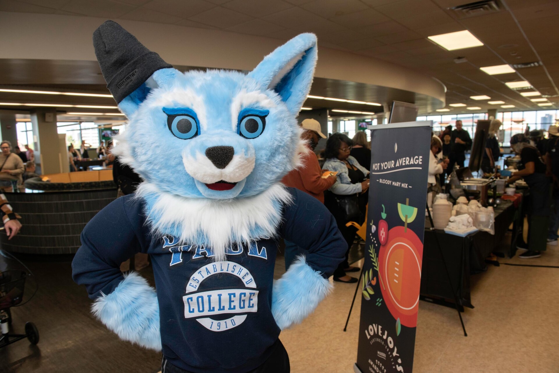 Sky the fox mascot wearing a Saint Paul College sweatshirt and a black hat stands with hands on hips in a busy indoor event space. Behind the mascot, people are gathered near tables with food and promotional signage.