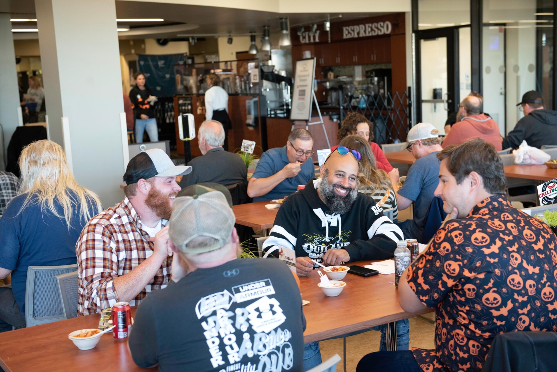 Groups of people sit at wooden tables in a cafeteria-style setting, eating bowls of chili. The background shows a coffee shop counter labeled “City Espresso” and additional diners